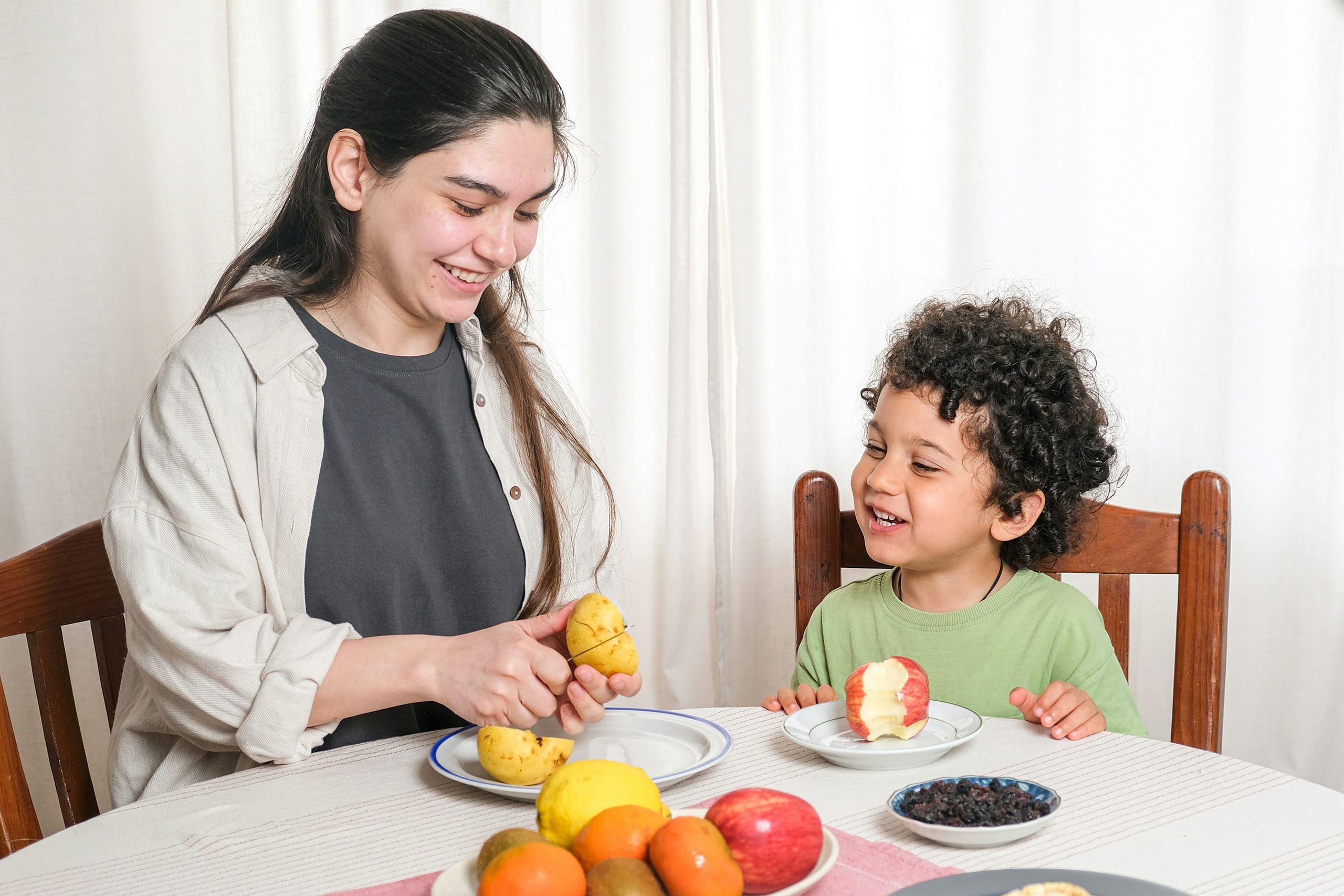 Nanny peeling fresh fruit for a young boy during snack time at the dining table