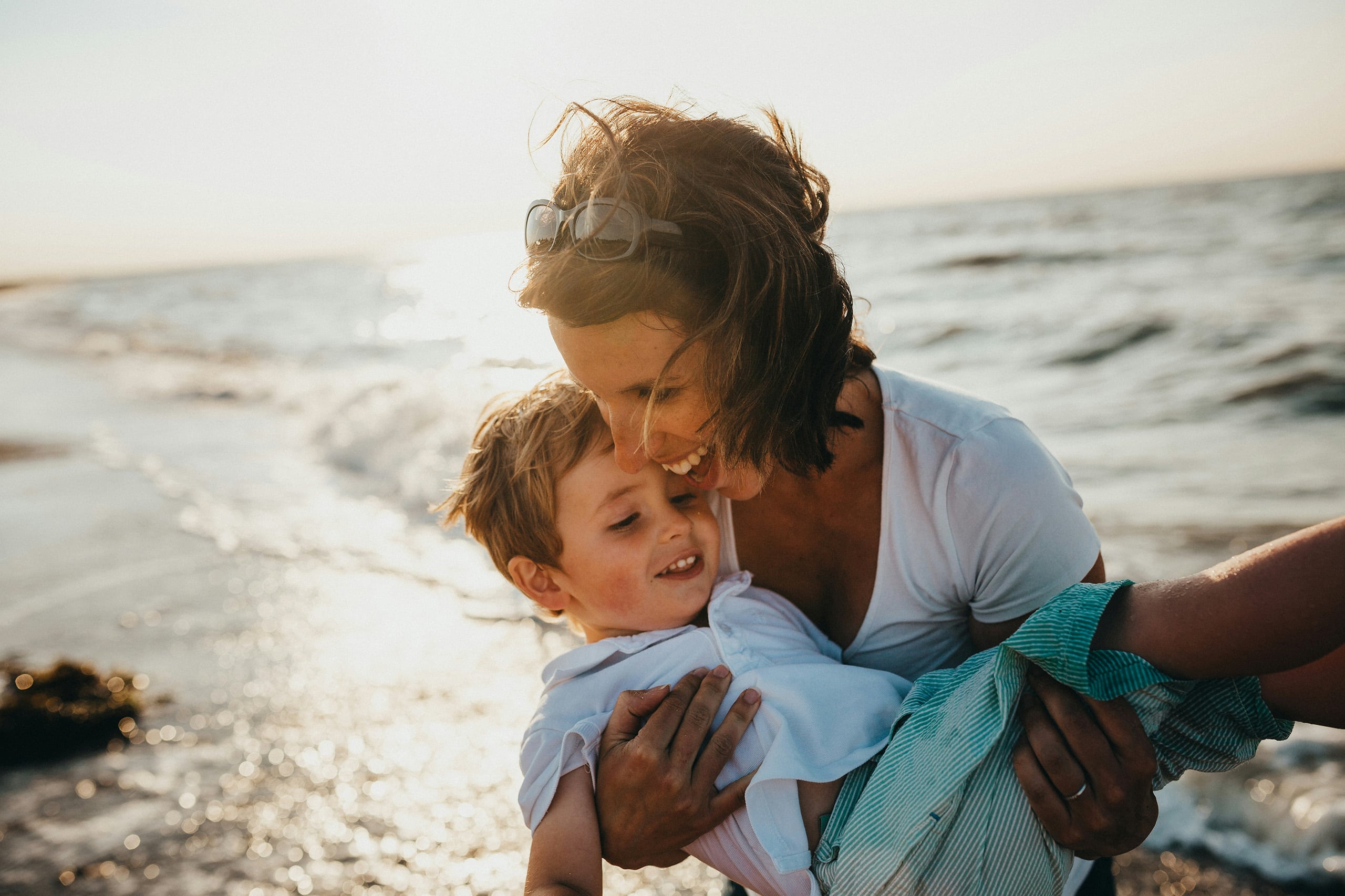 Travel nanny playfully holding a laughing young boy on a sandy beach during a family vacation