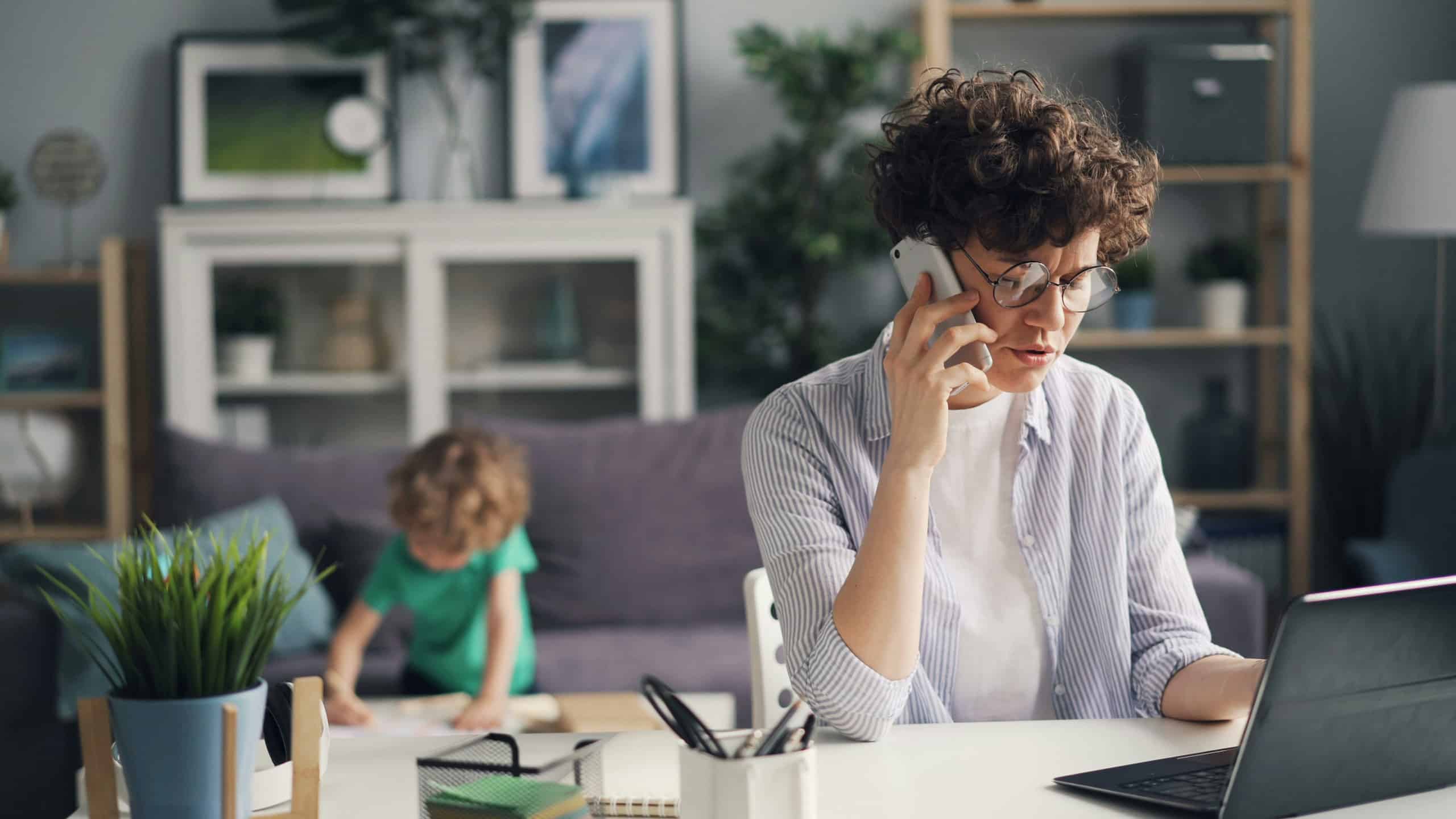 Working mother on a phone call while a reliable nanny plays with a child in the living room